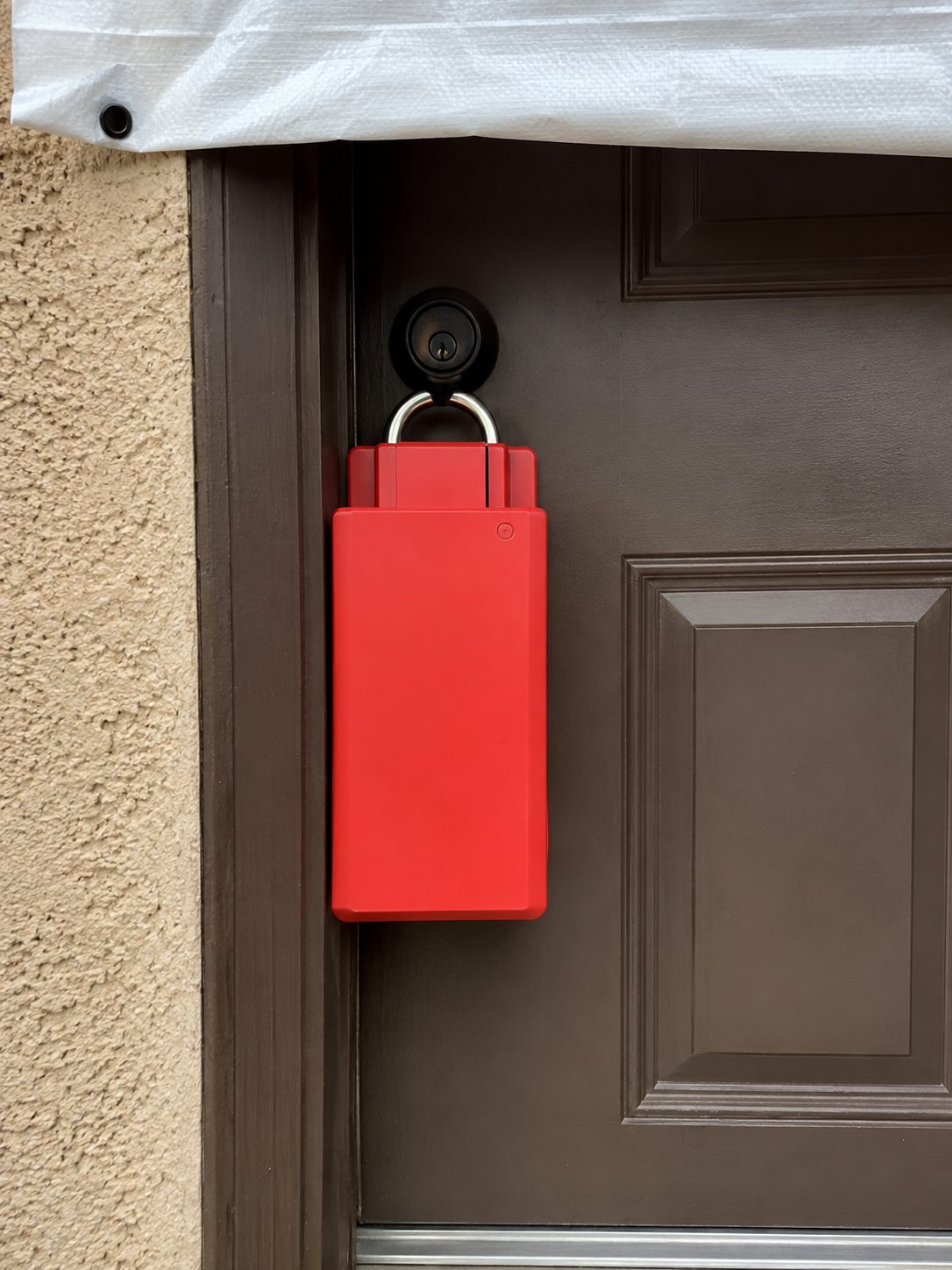 Heavy-duty red padlock lock-box installed over a residential front door's handle and deadbolt, with the fumigation tarpaulin visible above